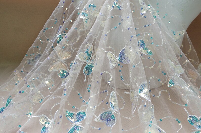 May include: White tulle fabric with iridescent blue and silver butterfly sequins. The fabric is sheer and delicate, with a soft, flowing texture.