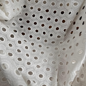 May include: Close-up of white eyelet fabric. The fabric features a repeating pattern of circular cutouts, each surrounded by delicate embroidered detailing. The fabric is slightly wrinkled, creating shadows and depth. The overall effect is a soft, textured, and airy appearance.