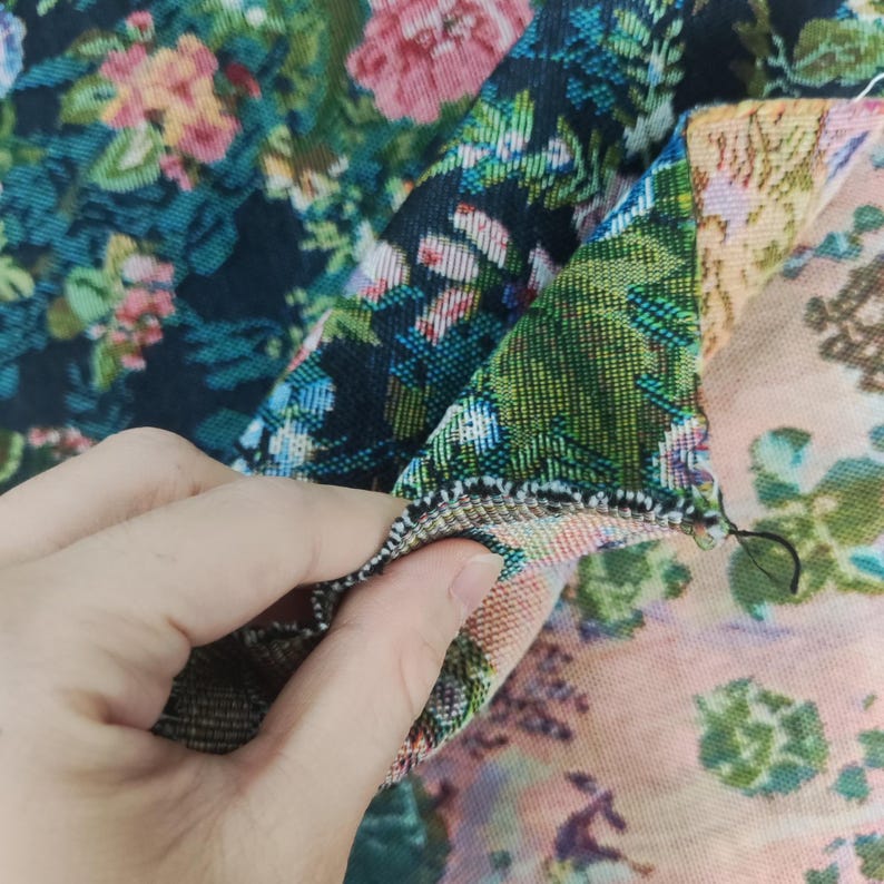 Puede incluir: Una tela tejida con un denso patr&oacute;n floral de flores rosas, verdes y azules sobre un fondo negro en un lado, mientras que el reverso muestra un dise&ntilde;o floral m&aacute;s claro en tonos melocot&oacute;n y verde con una costura visible.