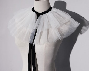 May include: A white tulle ruff collar featuring two layers of gathered, sheer fabric. The collar is secured around the neck with a black velvet ribbon that ties at the front, with two long, dangling black velvet ribbon ends hanging down.