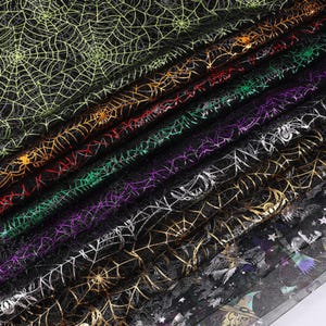 May include: A collection of folded black tulle fabric featuring metallic spiderweb patterns in various colours including lime green, orange, red, purple, silver, and gold, with one layer displaying a printed witch and star motif.