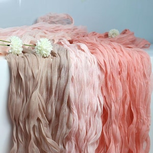 Puede incluir: Una exhibición de tela drapeada en tonos rosa y melocotón. La tela tiene una apariencia texturizada y fluida, con una calidad ligera y aireada. Acentos florales blancos se colocan encima de la tela.