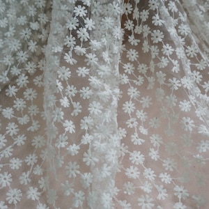 Può includere: Tessuto bianco trasparente con un delicato motivo floreale. Il tessuto è realizzato in una maglia fine e presenta piccoli fiori bianchi ricamati.