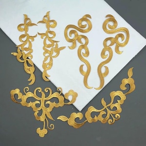 May include: A collection of six gold-colored embroidered appliques featuring intricate swirling and scrollwork patterns. These decorative patches are arranged in various shapes, including elongated vertical designs and wider, symmetrical motifs.