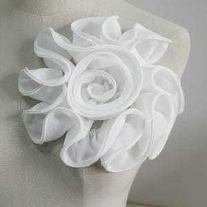 May include: A white fabric flower with layered, ruffled petals. The flower is designed in a spiral pattern, with the center tightly wound and the outer petals more open. The flower is likely a decorative accessory.