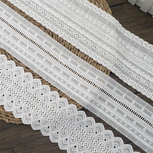 Puede incluir: Primer plano de tres cintas de encaje blanco con intrincados diseños florales y geométricos. El encaje está dispuesto diagonalmente sobre una superficie tejida, mostrando los delicados detalles. Ideal para costura y manualidades.