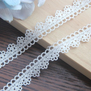 May include: White crochet lace trim with a delicate floral pattern. The lace is shown on a wooden surface.