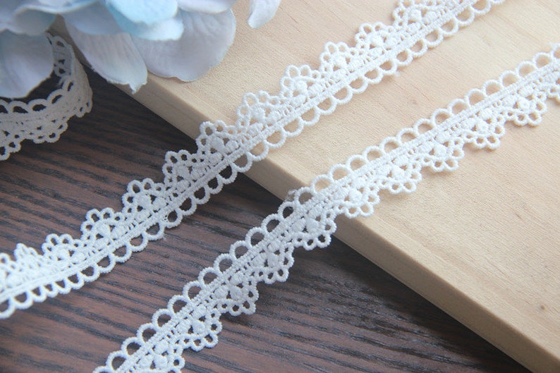 May include: White crochet lace trim with a delicate floral pattern. The lace is shown on a wooden surface.