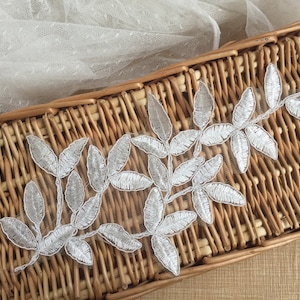 May include: Silver leaf embroidery trim with a delicate floral design. The trim is displayed on a woven basket.