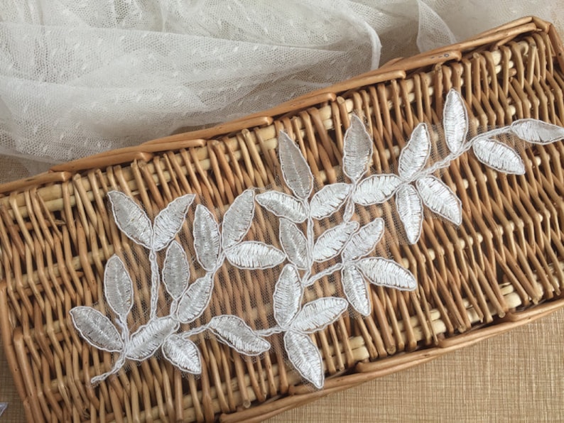 May include: Silver leaf embroidery trim with a delicate floral design. The trim is displayed on a woven basket.