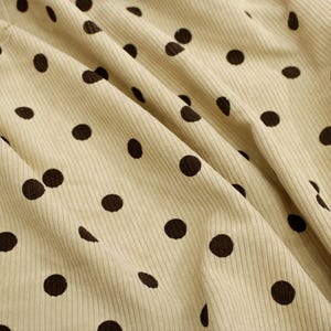 May include: A light beige corduroy fabric featuring a repeating pattern of dark brown polka dots. The material displays a distinct vertical ribbed texture with soft folds and drapes that create natural shadows across the surface of the textile.