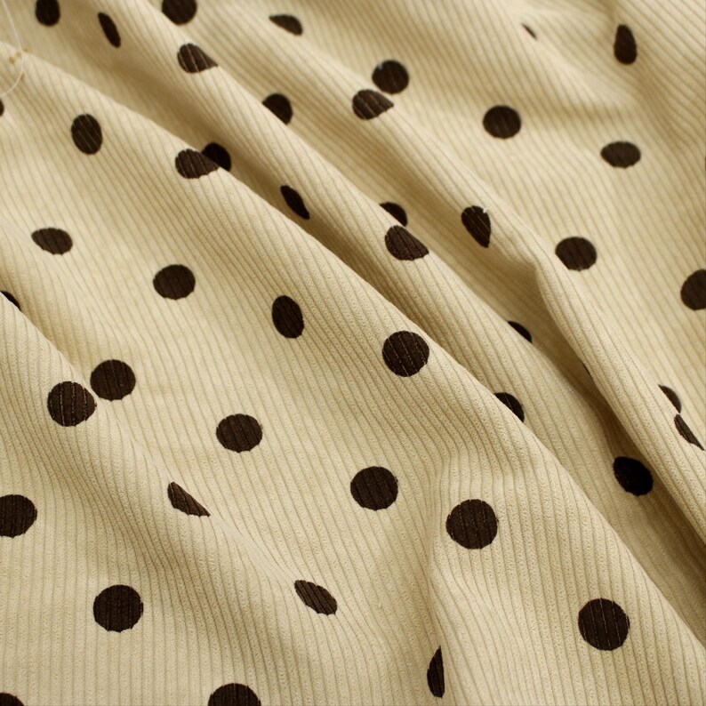 May include: A light beige corduroy fabric featuring a repeating pattern of dark brown polka dots. The material displays a distinct vertical ribbed texture with soft folds and drapes that create natural shadows across the surface of the textile.