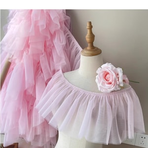 Puede incluir: Una blusa de tul rosa claro con diseño de volantes, exhibida en un maniquí. Una gran rosa rosa está prendida en la parte superior. En el fondo, un vestido rosa con volantes es parcialmente visible.