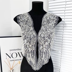 May include: A pair of white, beaded angel wing appliques on a black mannequin. The wings are made of a sheer, gray fabric and are heavily embellished with white beads and sequins. The text "yesterday NOW tomorrow" is visible in the background.