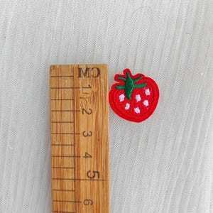 May include: A small, embroidered strawberry patch with a red body, green stem, and white seeds. A wooden ruler with centimeter markings is next to the patch. The patch is approximately 1 inch in size.