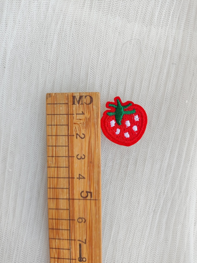 May include: A small, embroidered strawberry patch with a red body, green stem, and white seeds. A wooden ruler with centimeter markings is next to the patch. The patch is approximately 1 inch in size.