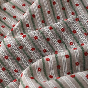 May include: Close-up of a fabric with a repeating pattern of red flowers and vertical stripes. The stripes alternate between white and green, set against a neutral, light brown background. The fabric is slightly wrinkled.
