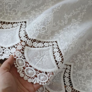 May include: Close-up of white lace fabric with intricate floral and geometric patterns. The fabric features detailed embroidery and a scalloped edge, showcasing a delicate and elegant design. The lace is held in a hand, highlighting the texture and craftsmanship.
