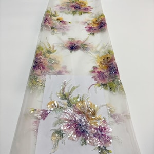 Puede incluir: Tela blanca transparente con bordados florales en tonos morados, amarillos y verdes. La tela presenta un patrón floral detallado con lentejuelas, ideal para ropa o decoración.