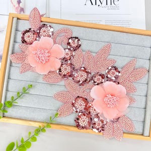 May include: A floral applique featuring two large, soft pink fabric flowers with layered petals and centers. The design includes numerous smaller, circular pink sequins and beaded leaves arranged in a sprawling, decorative pattern on a gray surface.