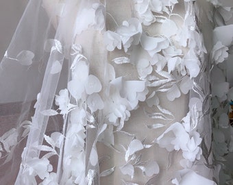 May include: White floral lace fabric with 3D flowers and leaves. The fabric is sheer and delicate, with a soft, romantic feel.