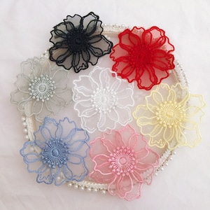 May include: A collection of seven embroidered flower appliques in black, red, white, light gray, pale yellow, light blue, and soft pink. Each flower features intricate thread detailing and a sheer, delicate structure arranged in a circular pattern.