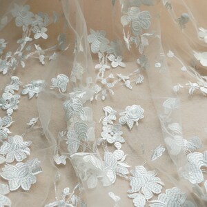May include: White floral lace fabric with a delicate pattern. The lace is sheer and has a soft, flowing texture.