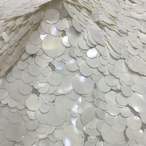 Puede incluir: Un tejido textil densamente cubierto de lentejuelas blancas circulares superpuestas de varios tama&ntilde;os. Las lentejuelas est&aacute;n dispuestas en un patr&oacute;n de capas, creando una superficie reflectante y texturizada con un acabado brillante que capta la luz.