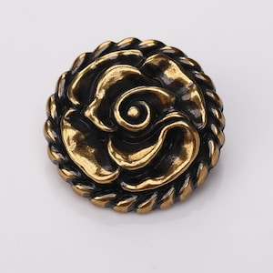 May include: A gold and black metal button featuring a raised rose design. The button has a rope-like border.