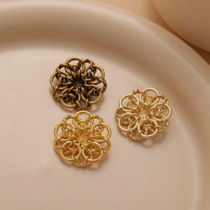 Puede incluir: Tres adornos de metal de color dorado en forma de flor con intrincados diseños circulares. Los adornos están dispuestos sobre una superficie de color marrón claro.