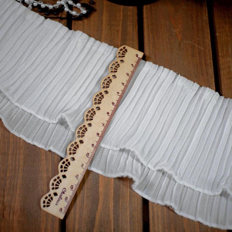 May include: A close-up of white pleated fabric with a decorative edge. A wooden ruler with a scalloped edge is placed diagonally across the fabric, showing centimeter measurements. The fabric appears to be a trim or edging material.