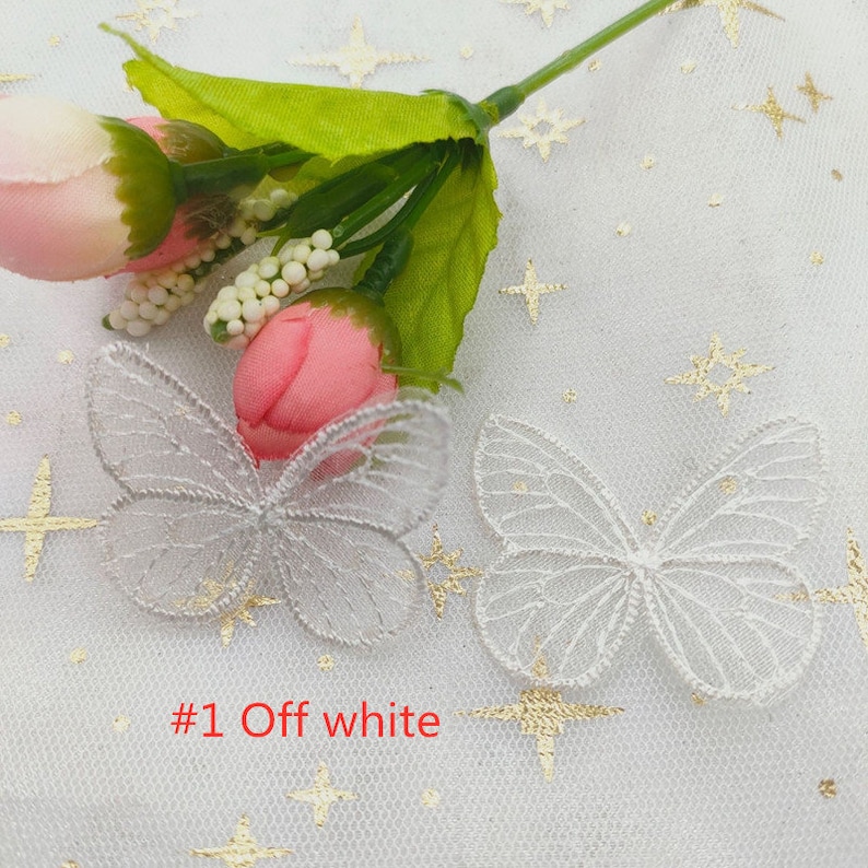 May include: Two white lace butterfly appliques with a gold glitter finish on a white background with gold glitter stars. The text #1 Off white is visible in the image.