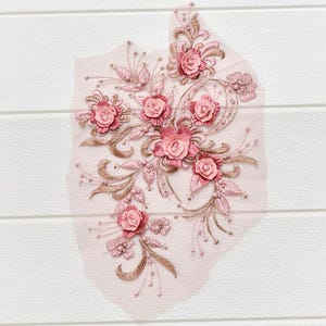 May include: A delicate, floral applique featuring pink and rose-colored fabric flowers, leaves, and beaded accents. The design is embroidered on a sheer, light pink mesh backing, creating a layered effect. The applique is ideal for embellishing garments or craft projects.