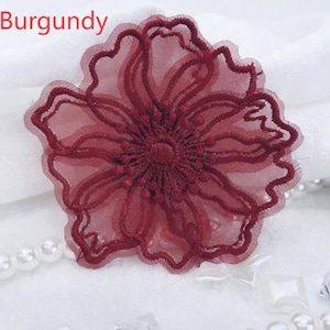 May include: A burgundy flower applique featuring sheer mesh fabric with intricate embroidered outlines and a textured center. The word Burgundy is printed in red text in the upper left corner of the image, which also includes white pearl beads.