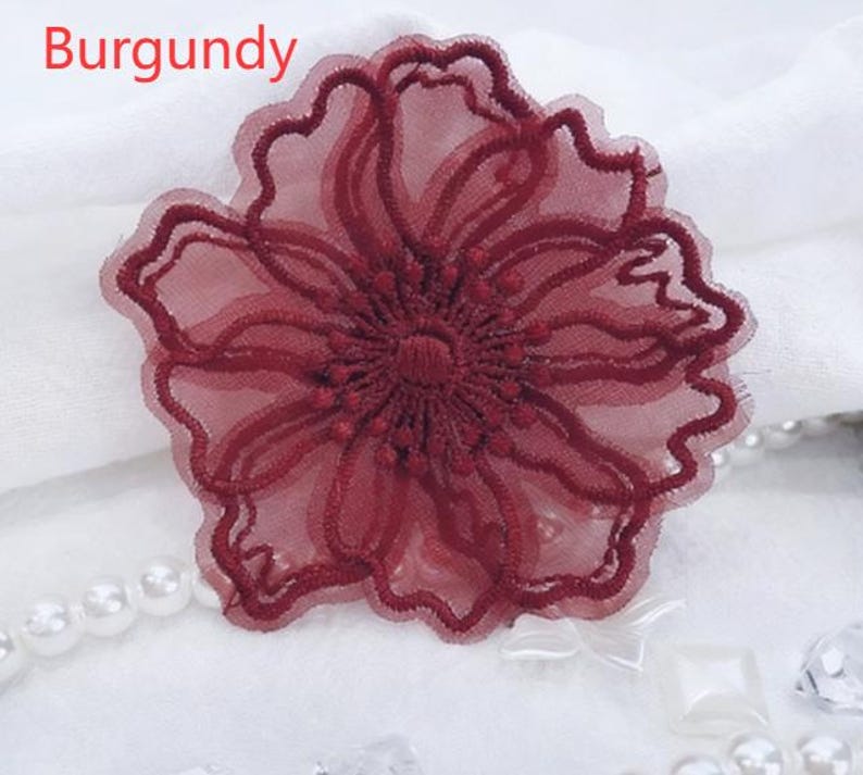 May include: A burgundy flower applique featuring sheer mesh fabric with intricate embroidered outlines and a textured center. The word Burgundy is printed in red text in the upper left corner of the image, which also includes white pearl beads.