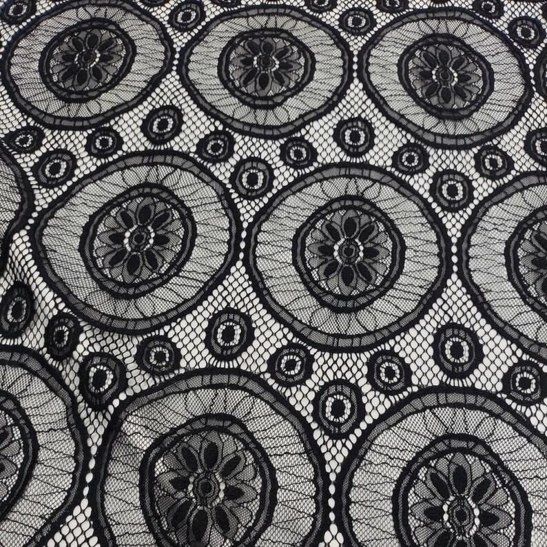 Peut inclure: Un tissu en dentelle noire pr&eacute;sentant un motif r&eacute;p&eacute;titif de grands cercles avec des centres floraux complexes. Le design comprend de plus petits accents circulaires intercal&eacute;s entre les grands cercles, reli&eacute;s par une fine maille ajour&eacute;e.