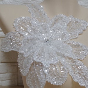 May include: A white floral applique with sequins and a small rhinestone in the center. The applique is made of delicate lace and is perfect for adding a touch of elegance to any garment.