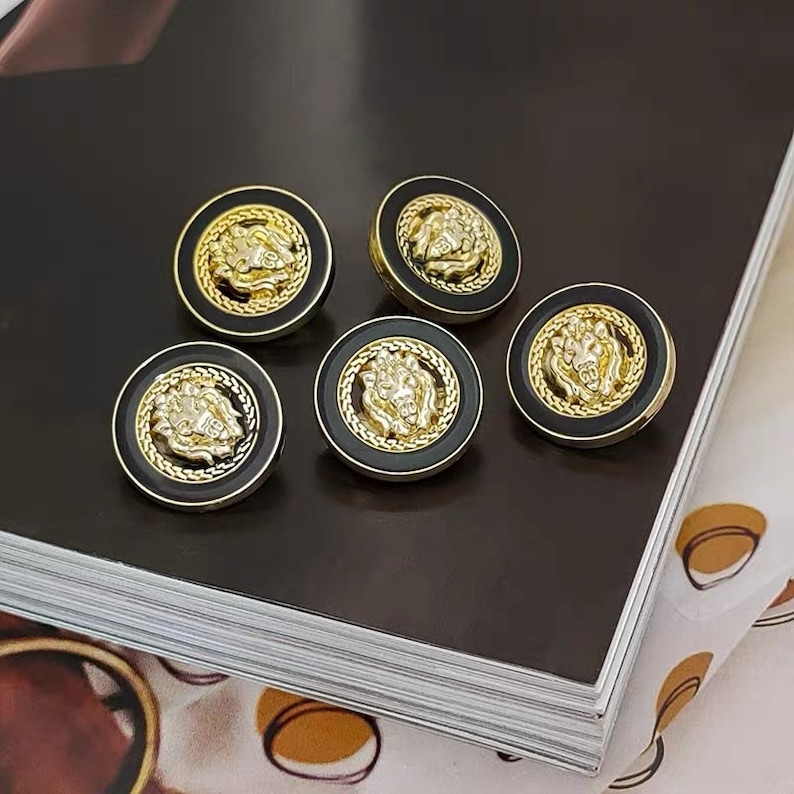 Puede incluir: Cuatro botones redondos dorados y negros con un dise&ntilde;o de cabeza de le&oacute;n en el centro. Los botones tienen un fondo negro y un borde dorado con un patr&oacute;n decorativo.
