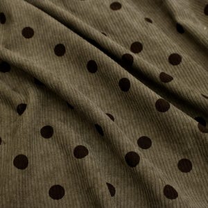 May include: An olive green corduroy fabric featuring a repeating pattern of dark brown polka dots. The material has a distinct vertical ribbed texture characteristic of corduroy, with soft folds creating depth and shadows across the surface.