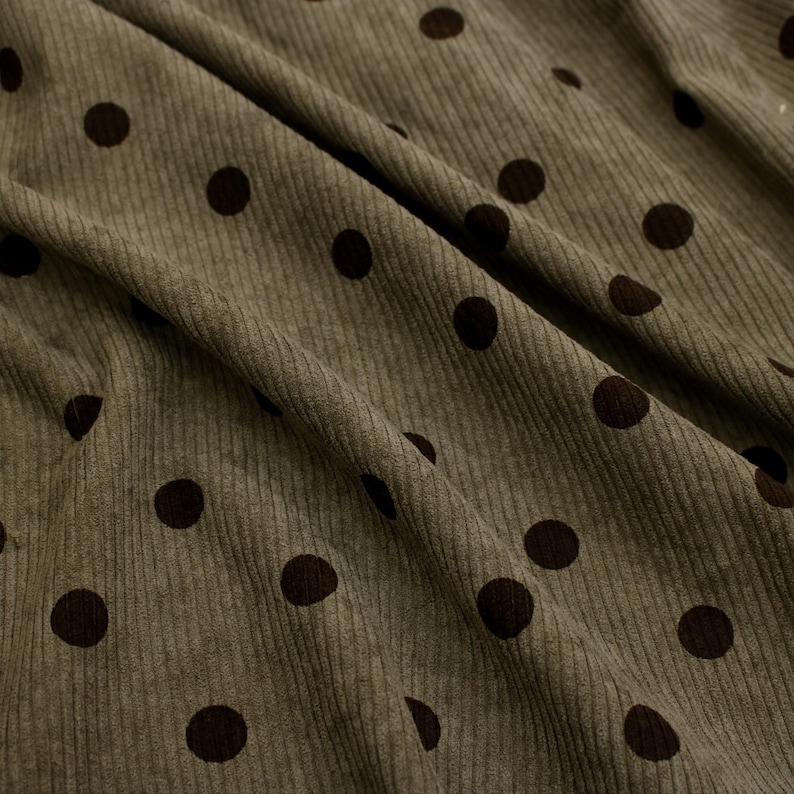 May include: An olive green corduroy fabric featuring a repeating pattern of dark brown polka dots. The material has a distinct vertical ribbed texture characteristic of corduroy, with soft folds creating depth and shadows across the surface.