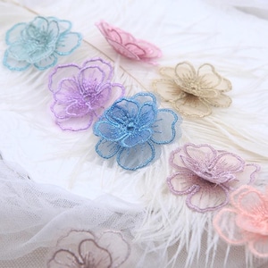 May include: A collection of multi-colored floral appliques made from sheer, embroidered mesh fabric. The flowers feature layered petals in shades of light blue, lavender, pink, gold, and peach, each with a delicate stitched outline and center.