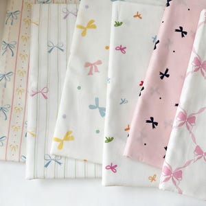 Op de afbeelding: Een verzameling gevouwen stofstalen met strikpatronen in pasteltinten. De stoffen tonen roze, blauwe, gele en groene strikken op witte en lichtroze achtergronden. Sommige hebben gestreepte of gestippelde patronen.
