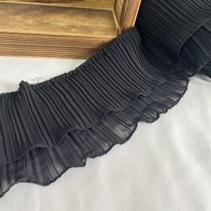 May include: Close-up of a length of black pleated fabric. The fabric is layered, with a ruffled edge. The material appears to be lightweight and semi-sheer. The fabric is resting on a white surface, with a wooden box in the background.