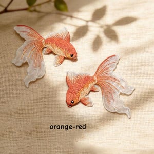 May include: Two embroidered goldfish patches featuring orange-red bodies and translucent white flowing fins. The fish are depicted in a swimming motion with detailed stitching. The text orange-red is printed below the patches.