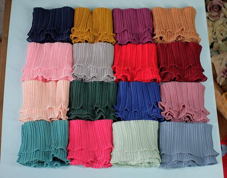 May include: A collection of pleated fabric pieces in various colors, including navy, yellow, pink, gray, red, and teal. Each piece has a ruffled edge, creating a layered effect. The fabric appears to be lightweight and suitable for clothing or craft projects.