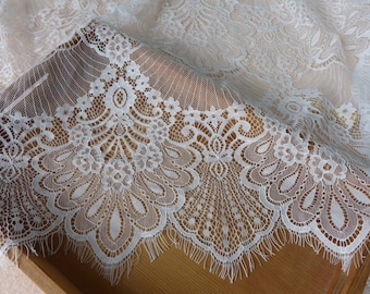 May include: Close-up of white lace fabric with a detailed floral pattern. The lace features a scalloped edge with delicate fringe. The fabric is draped over a wooden surface, showcasing the intricate design and texture of the lace.