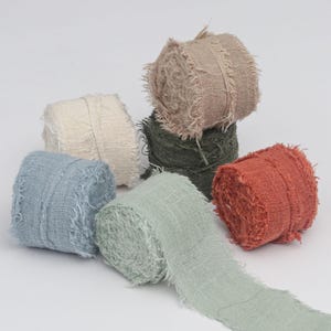 May include: A collection of six spools of frayed-edge fabric ribbon in various colors including cream, light blue, sage green, dark olive green, terracotta orange, and beige, arranged in a cluster with one spool partially unrolled.