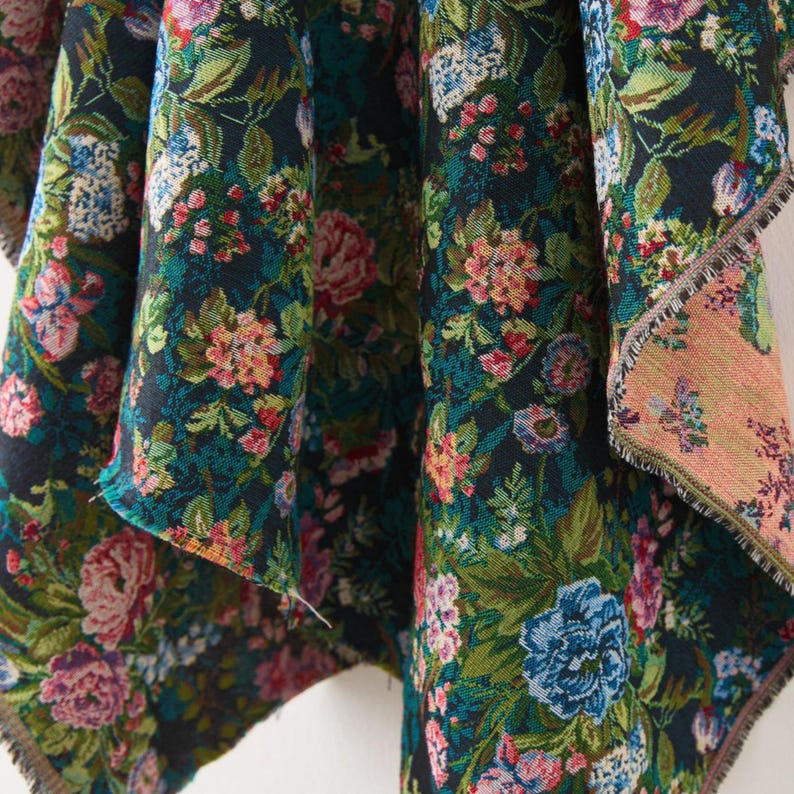 Puede incluir: Una tela tejida con un fondo oscuro y un denso patr&oacute;n floral en tonos rosa, azul, verde y blanco. El textil muestra un tejido complejo estilo tapiz con bordes deshilachados visibles y un reverso de color melocot&oacute;n claro que contrasta.