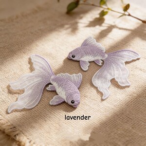 May include: Two lavender embroidered fish patches featuring intricate thread work and flowing fins. The patches are shaped like goldfish with detailed scales and delicate, semi-transparent fin edges. The word lavender is printed below the fish.