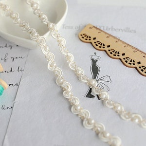 May include: White beaded trim with a scalloped edge. The trim is made of small white pearls and a delicate lace pattern.